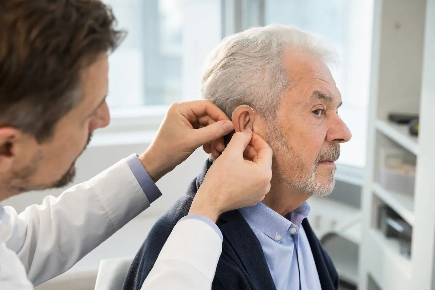 Un audioprothésiste ajuste une aide auditive de classe 1 derrière l'oreille d'un patient senior dans son cabinet