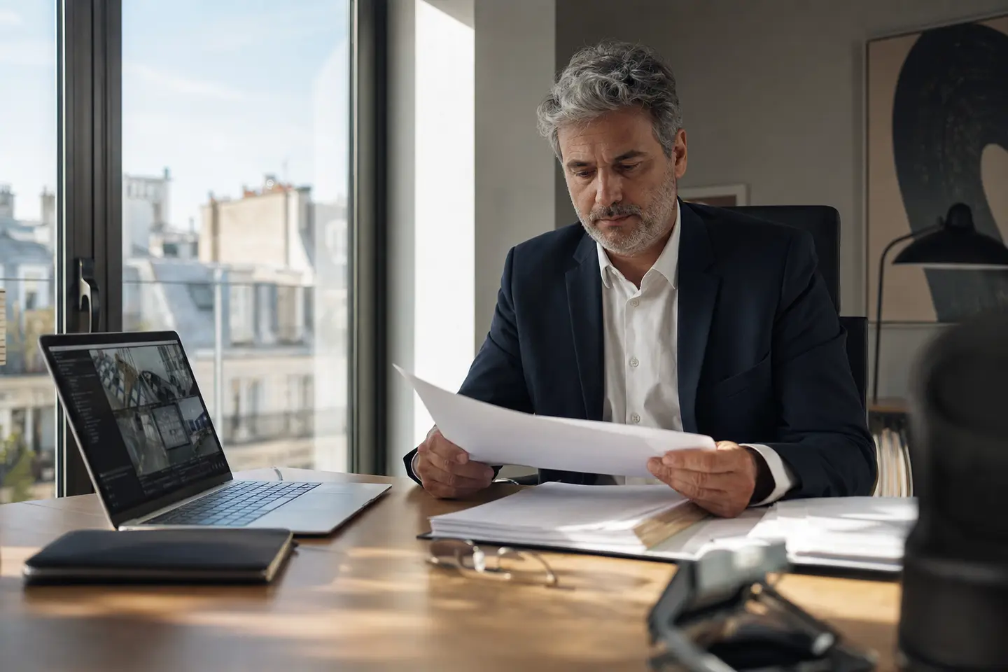 Dirigeant d’entreprise française lisant un contrat à son bureau, tableau de bord de sécurité informatique ouvert sur son ordinateur portable