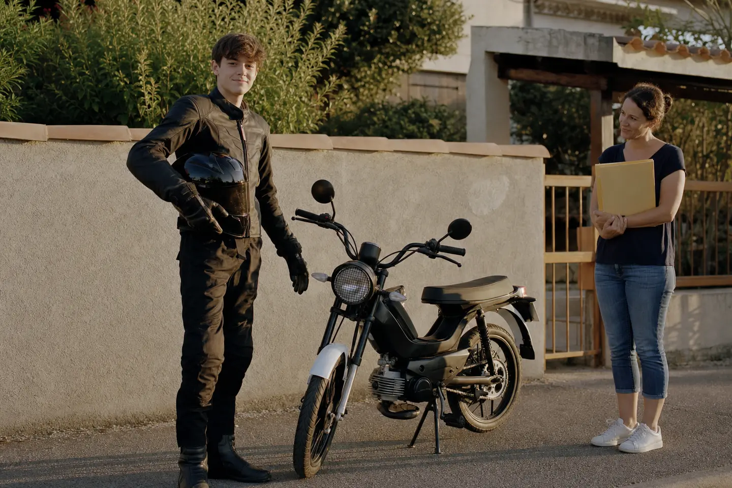 Adolescent de 16 ans en équipement moto à côté de sa 125 cm³ accompagné d'un parent tenant un dossier