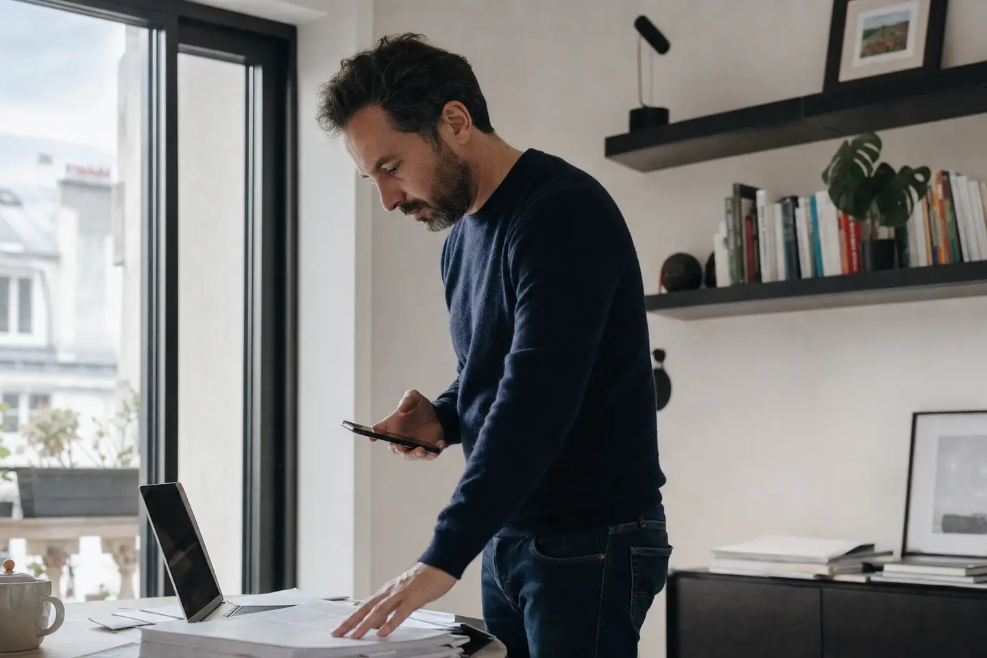 Homme debout près d'une fenêtre consultant des documents après une perte d'emploi dans un bureau à domicile.