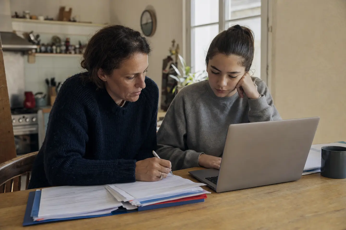 Un parent et un adolescent remplissant des documents à la table de la cuisine, dossier de preuves et ordinateur ouverts, en vue d'un dépôt de plainte