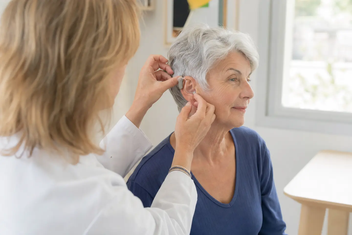 Une audioprothésiste ajuste une aide auditive discrète sur l'oreille d'une patiente senior en cabinet